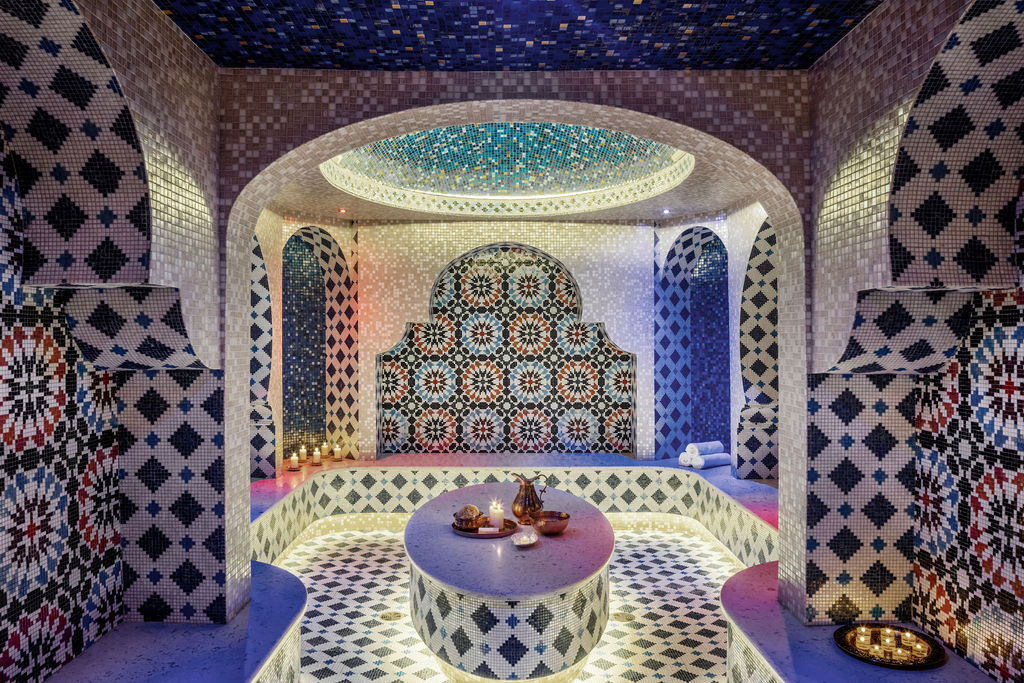 A Moroccan hammam spa room bathed in soft purple and blue light. The walls and arches are adorned with intricate mosaic tiles in patterns of white, black, and red. A central circular bench covered in small tiles creates a focal point, with spa essentials set atop. Above, the ceiling features a large, illuminated oval recess, contributing to the room's serene and exotic ambiance.