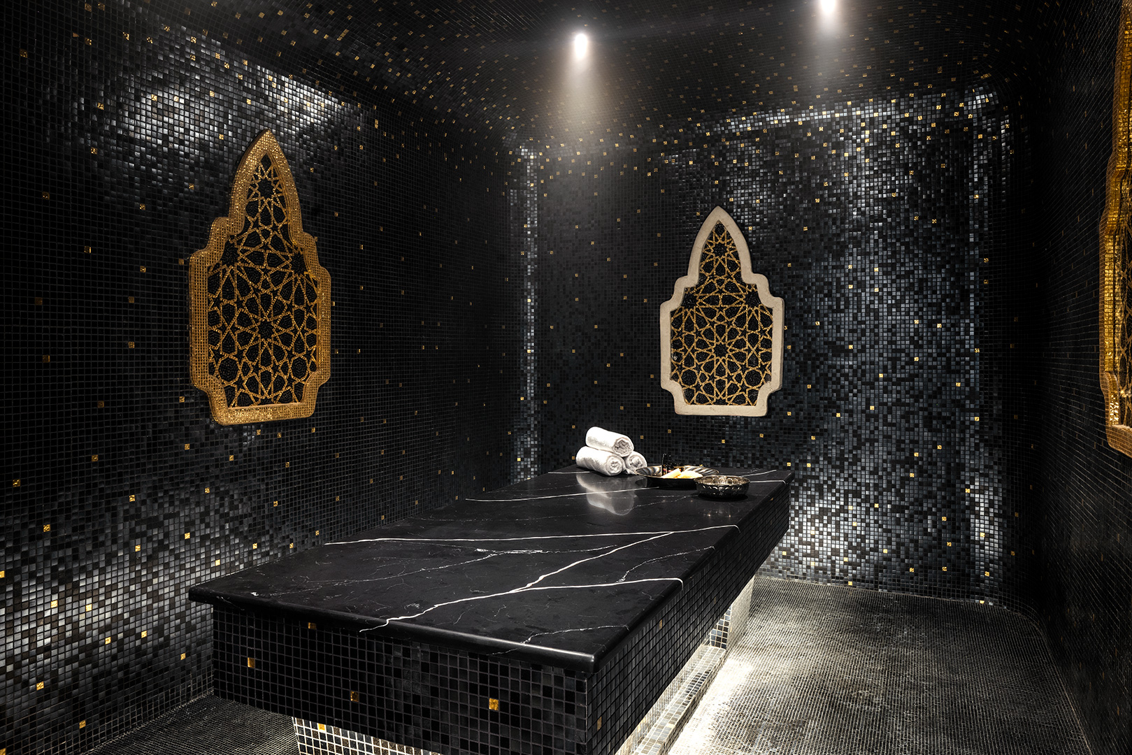 A luxurious private hammam room adorned with black mosaic tiles and gold accents. Two ornate, gold-framed wall features add a touch of elegance. The centerpiece is a large, black marble slab for reclining, with white towels and spa essentials neatly arranged to one side, all illuminated by soft ceiling lights.