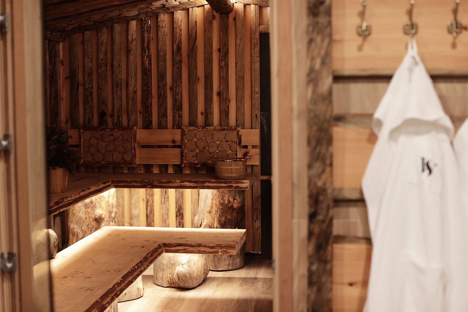 An empty sauna with rustic wooden benches and walls, complete with traditional sauna accessories like a wooden bucket and ladle. A white robe with a monogram WS is hanging on the wall, ready for use.