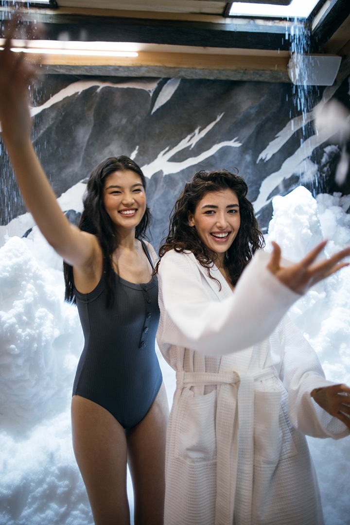 Two people are joyfully interacting with a snow in a snow room with a mountainous mural and soft lighting, creating a playful indoor winter scene.