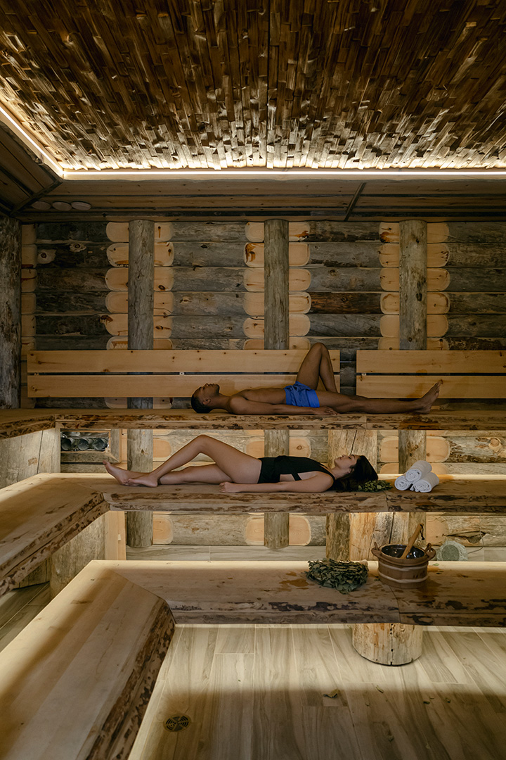 This image features two individuals relaxing in a traditional wooden sauna. The room is constructed with natural logs, creating a cabin-like atmosphere. The benches are arranged in stepped tiers allowing for different levels of heat. On the lowest bench, there's a wooden bucket and a ladle, essential for a sauna experience. The ceiling is adorned with cross-sections of logs, and gentle light filters through the room, accentuating the serene ambiance.