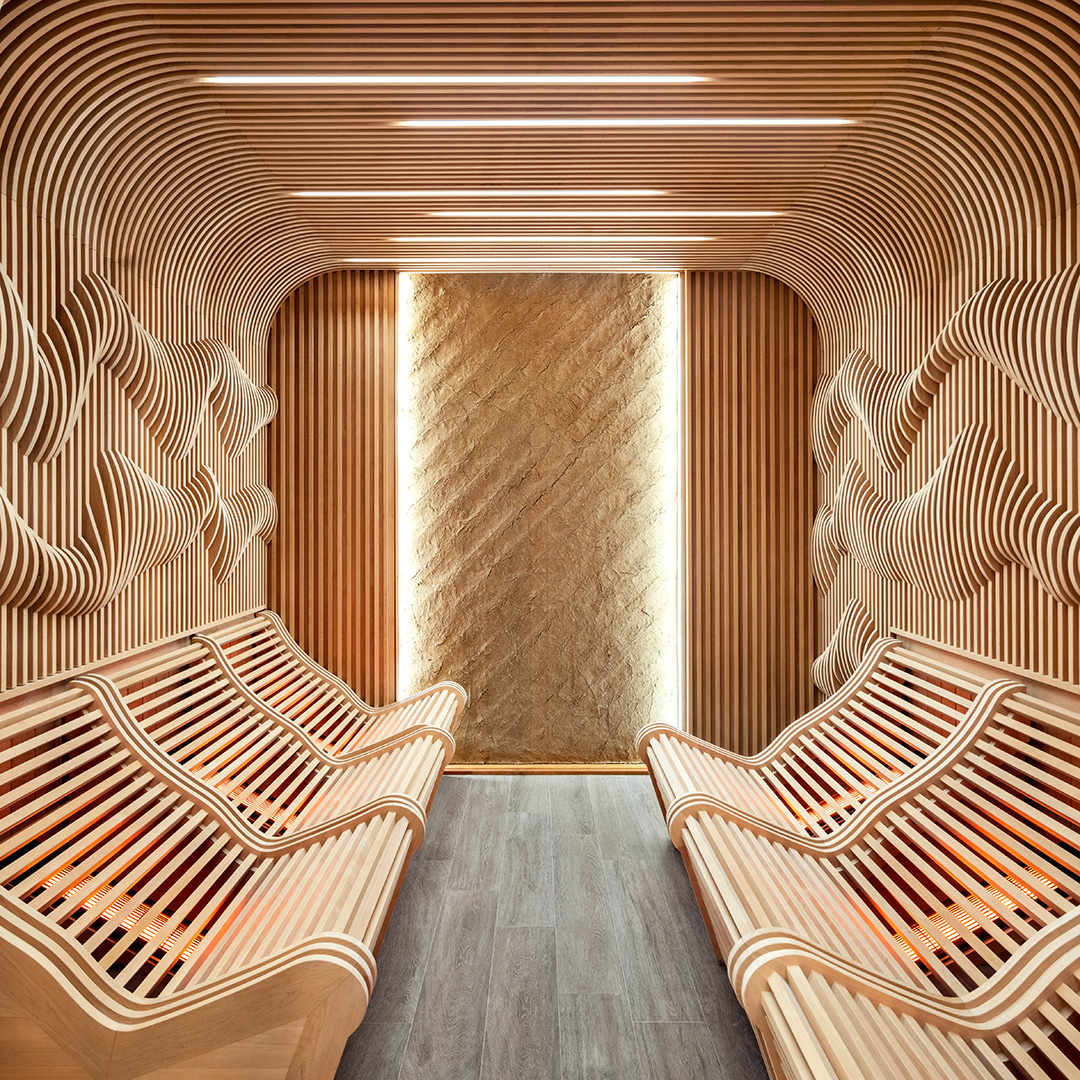A modern sauna with a unique design featuring curved wooden slats along the walls and ceiling, creating a wave-like pattern. Ergonomically shaped benches with built-in heating elements offer a comfortable seating experience. The room is softly lit, emphasizing the wood's natural grain, enhancing the relaxing and contemporary atmosphere.