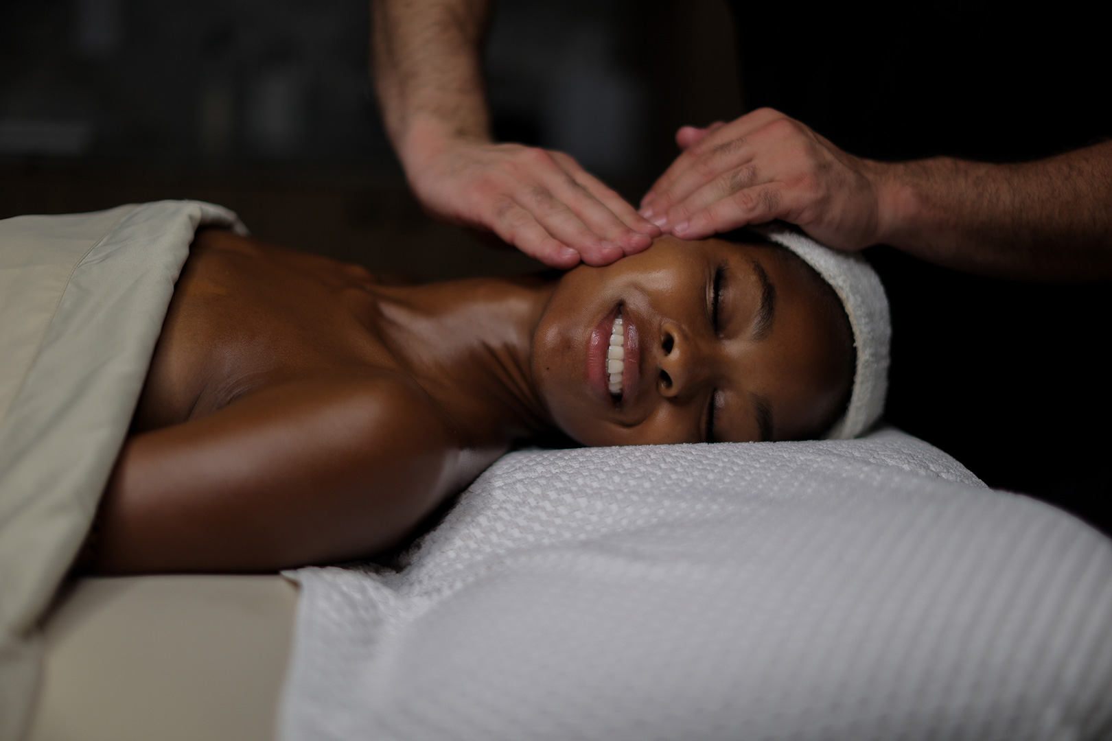 A person is lying down, appearing relaxed with a serene expression, receiving a head massage. They are draped in a white spa towel, emphasizing a calm and therapeutic setting.