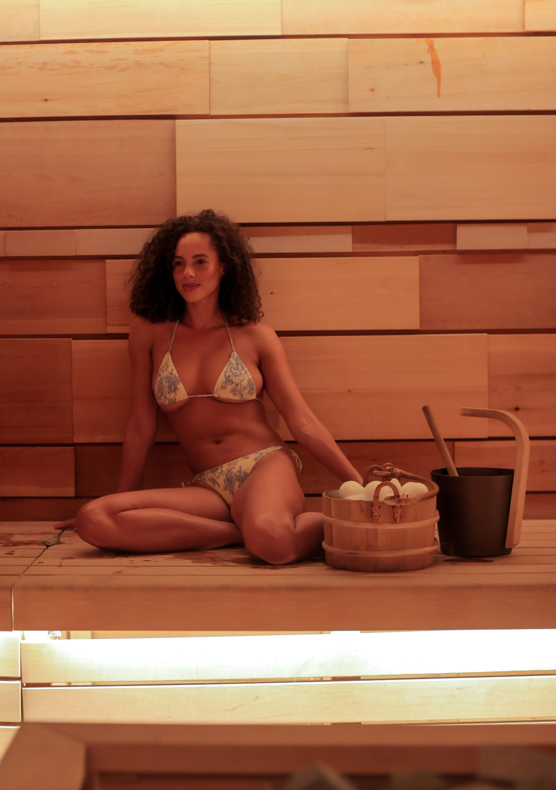 A person is seated in a sauna with wooden walls and benches, and items for a sauna session nearby. The atmosphere is warm and tranquil.