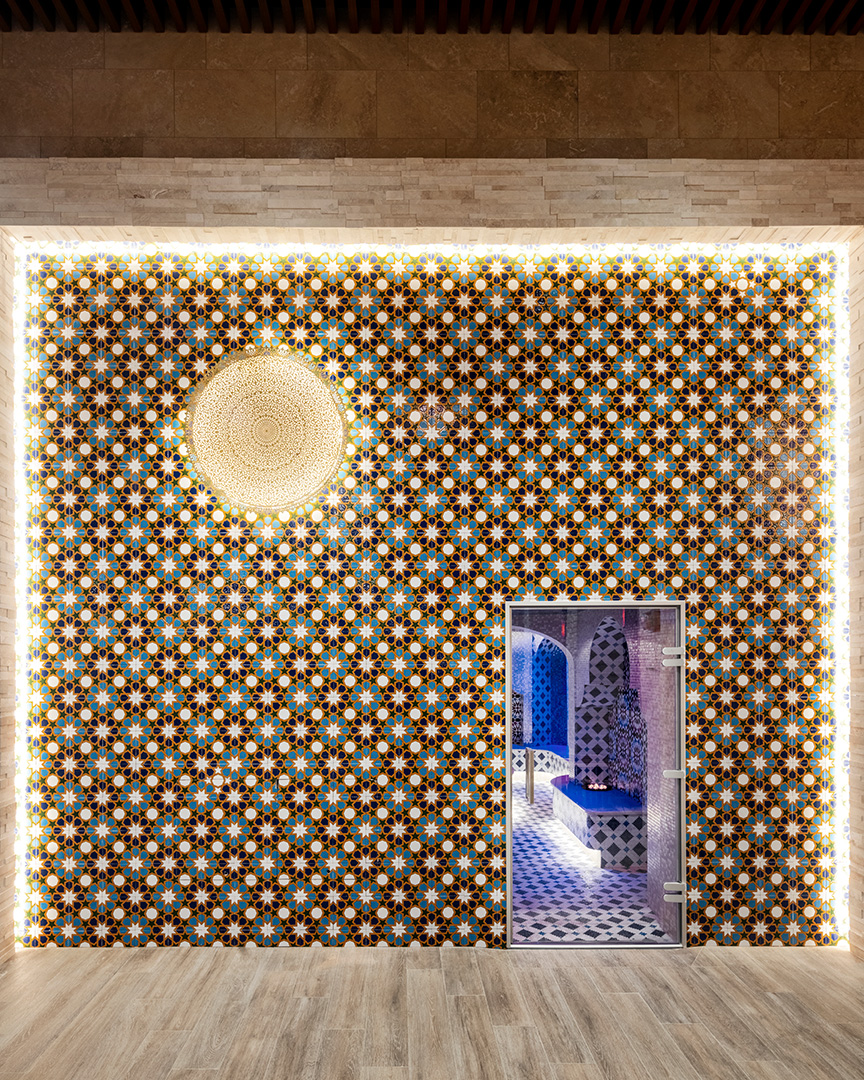 A wall of marroccan hammam room with a patterned wall and an open door. The wall is ornate with blue, white, and gold geometric shapes. A golden light fixture hangs in the center. The visible floor is wooden.