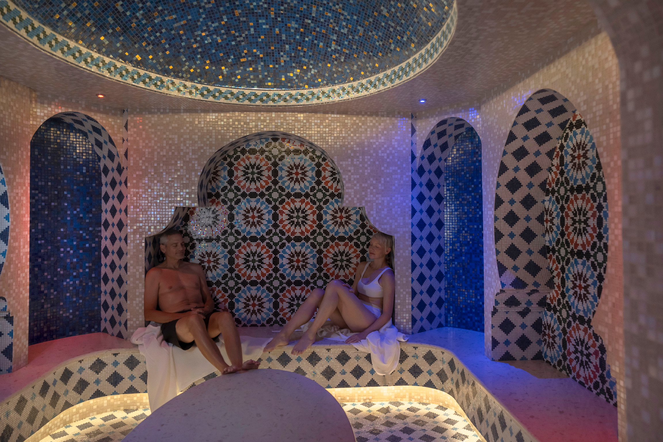 Two individuals relaxing in a colorful tiled morroccan hammam room.