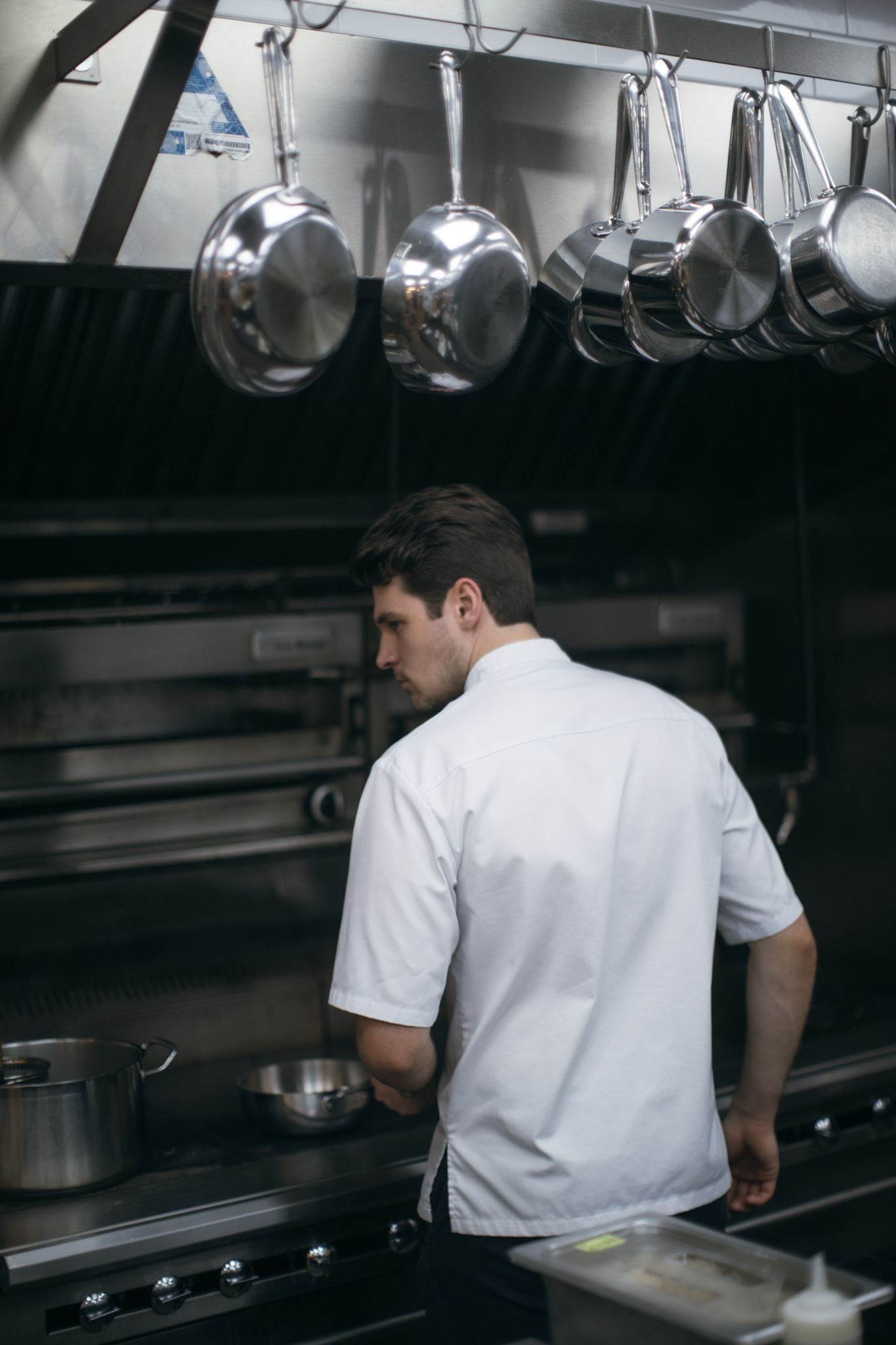 A person in a white chef's uniform is working in a commercial kitchen, standing in front of a stainless steel stove with pots and pans. The kitchen is equipped with hanging cookware overhead, indicating a professional cooking environment