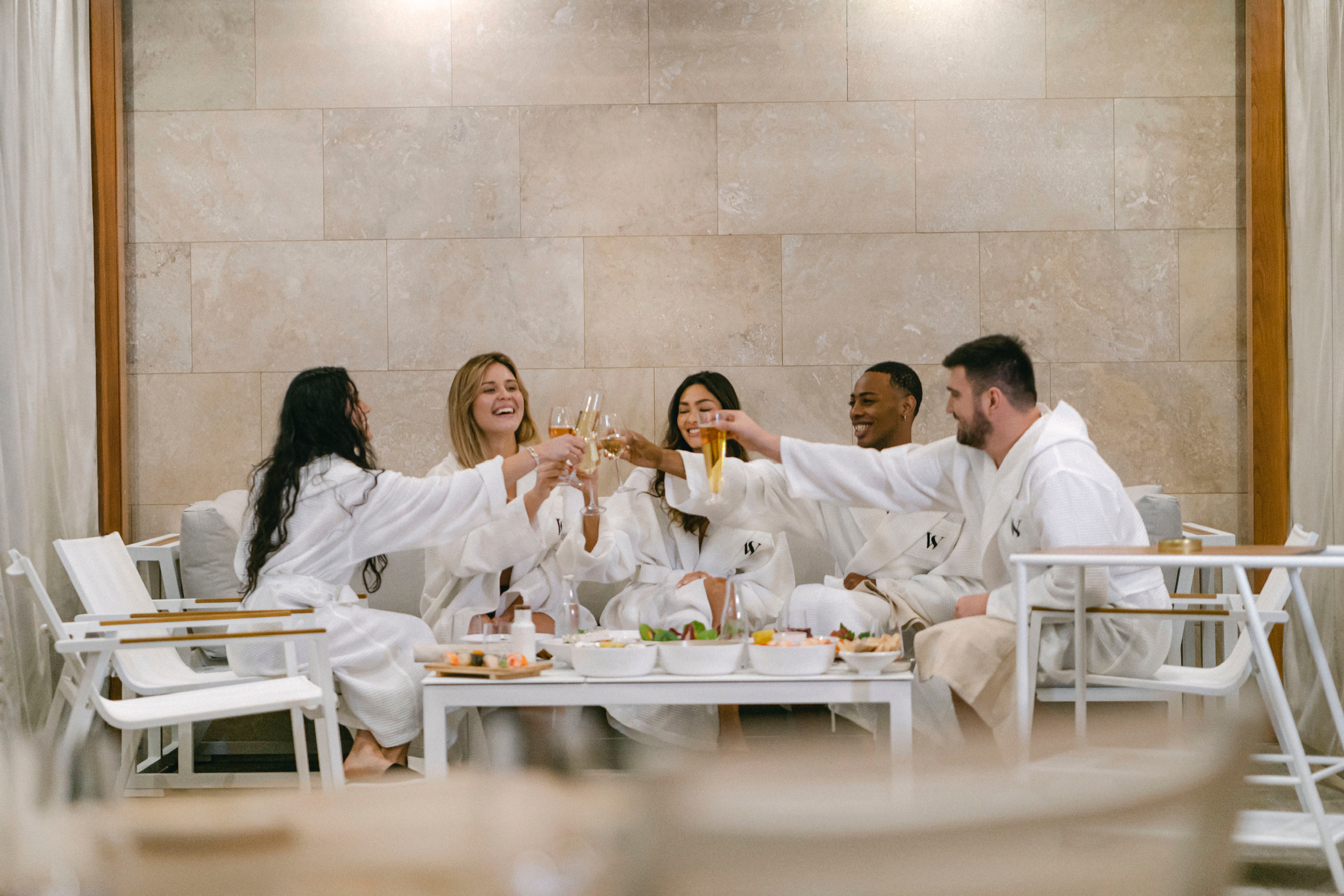 A group of people in white robes are toasting with glasses, smiling and enjoying a social moment around a low table with light refreshments, in a room with tiled walls and a relaxed spa ambiance.