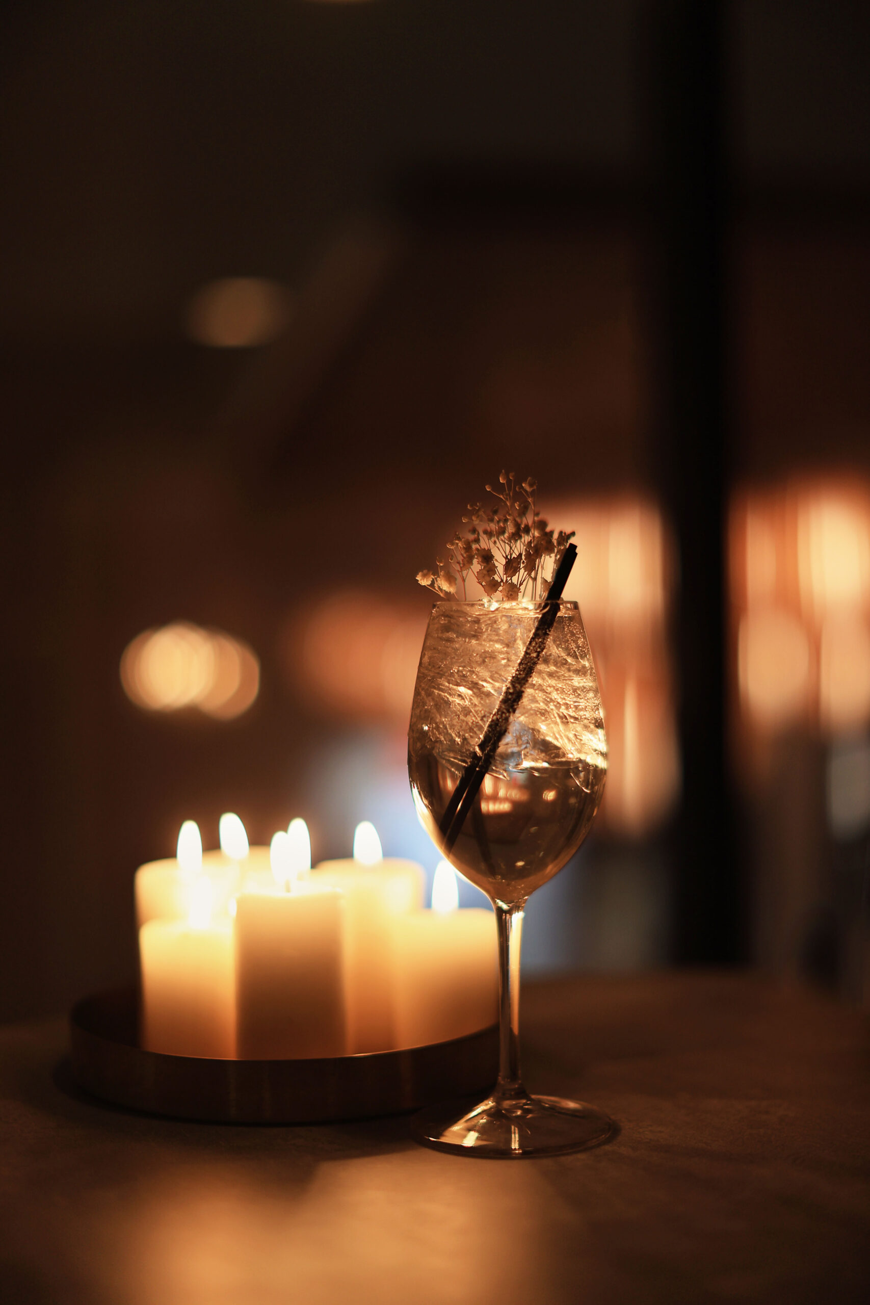 A cocktail with a straw and decorative herbs set next to a tray of lit candles.