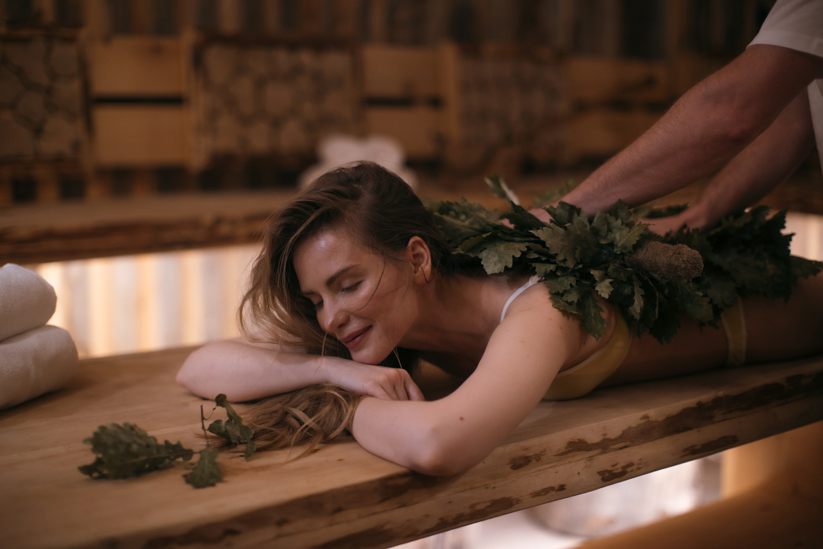 A person is lying face down on a wooden bench in a sauna, looking relaxed and serene. They are receiving a traditional sauna treatment with leafy branches being gently brushed along their back by another individual