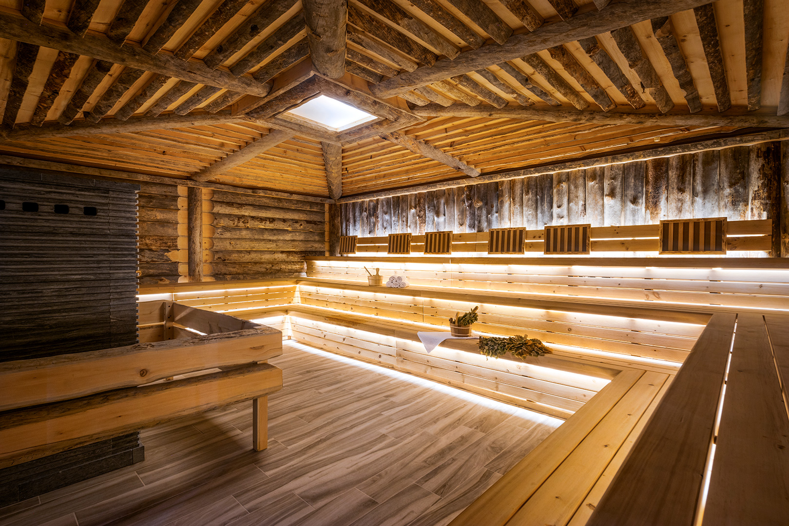 This image shows the interior of a traditional sauna room. The space is characterized by its rustic charm, with walls and ceilings made of raw, exposed logs creating a cabin-like feel. Wooden benches are arranged in tiers along the walls, providing seating. There's a soft, natural light coming through a skylight in the center of the ceiling, and LED strips under the benches add a modern touch. A sauna heater made of stacked stones is situated in one corner, and there are wooden buckets and ladles, likely for pouring water on the hot stones to create steam.
