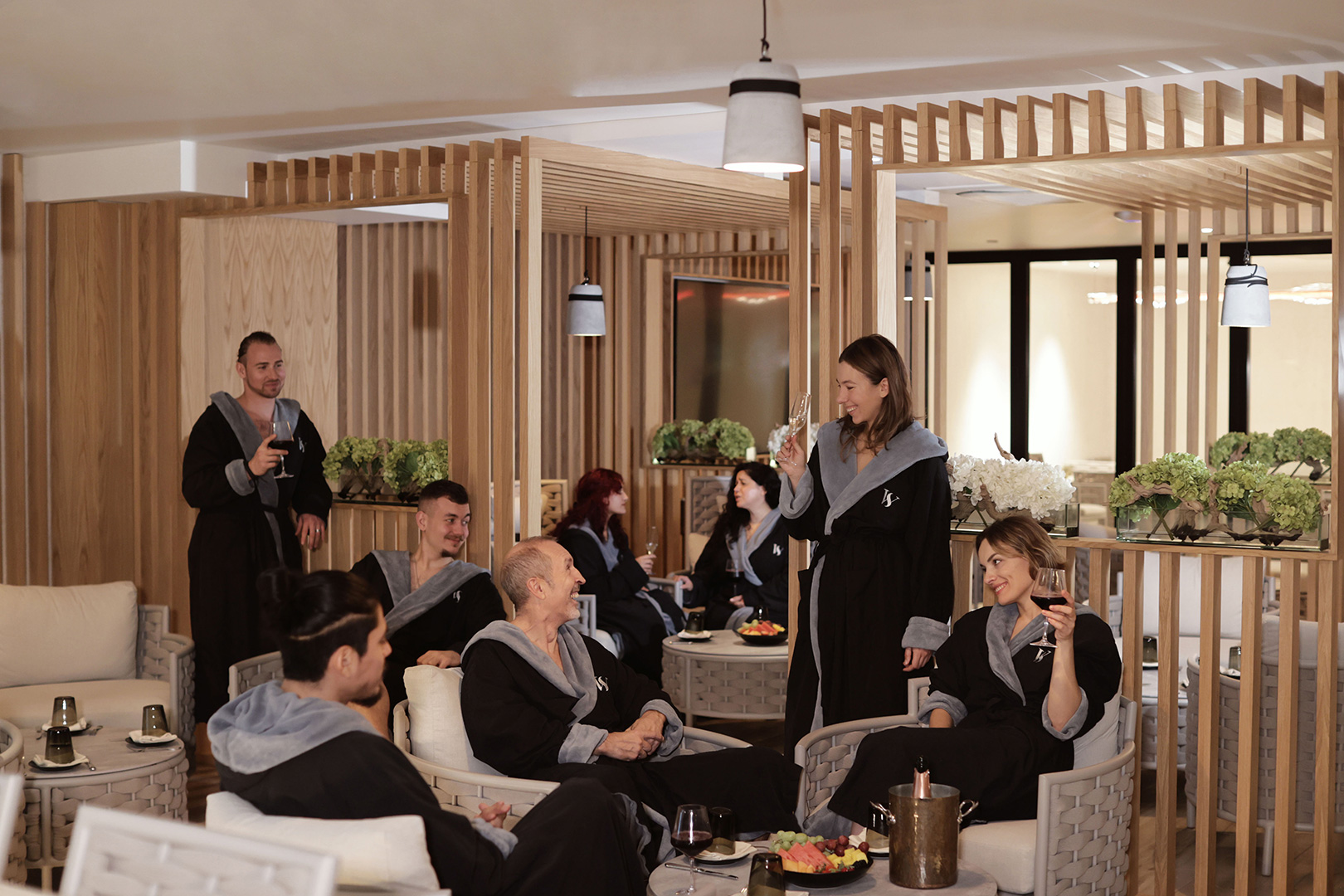 A lively social gathering in a luxurious lounge area, with guests enjoying themselves in a relaxed setting. The room features contemporary decor with plush seating and elegant wooden partitions. Attendees are dressed in spa robes, suggesting a casual yet exclusive event, with waitstaff providing attentive service.