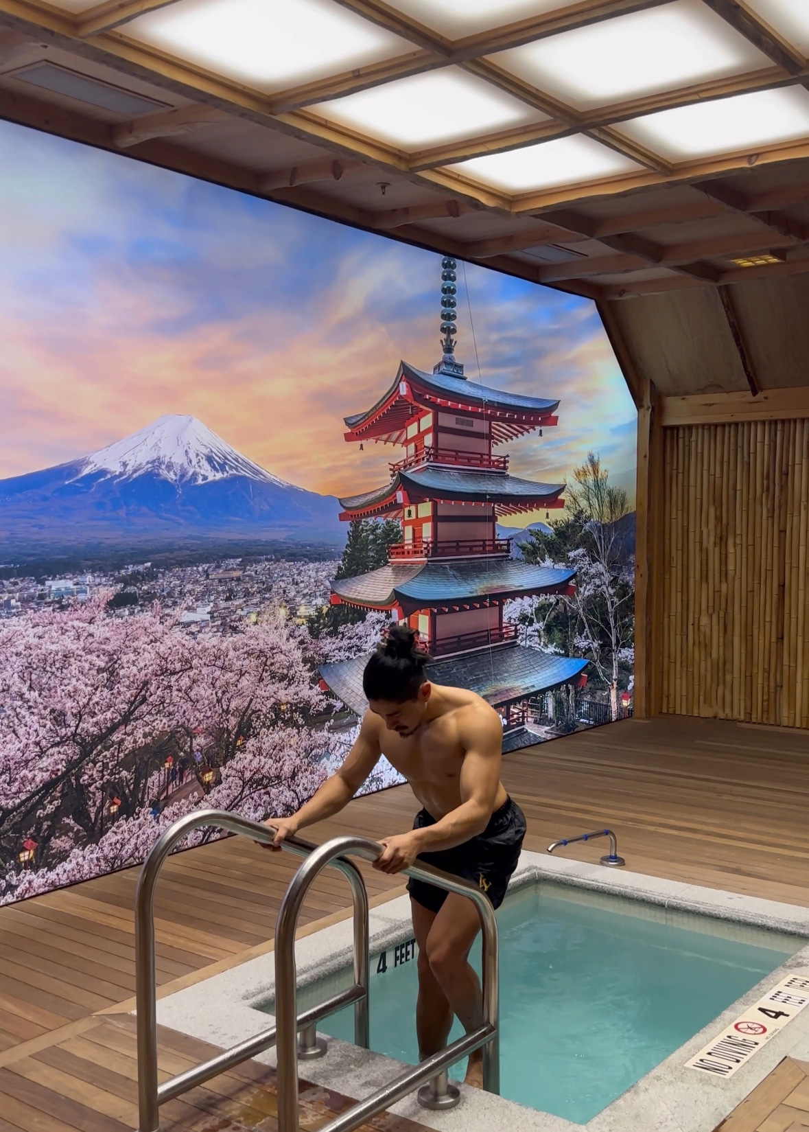 A person is stepping into a hot tub in a spa with a large mural of Mount Fuji and a pagoda surrounded by cherry blossoms in the background, creating a serene Japanese-inspired setting.