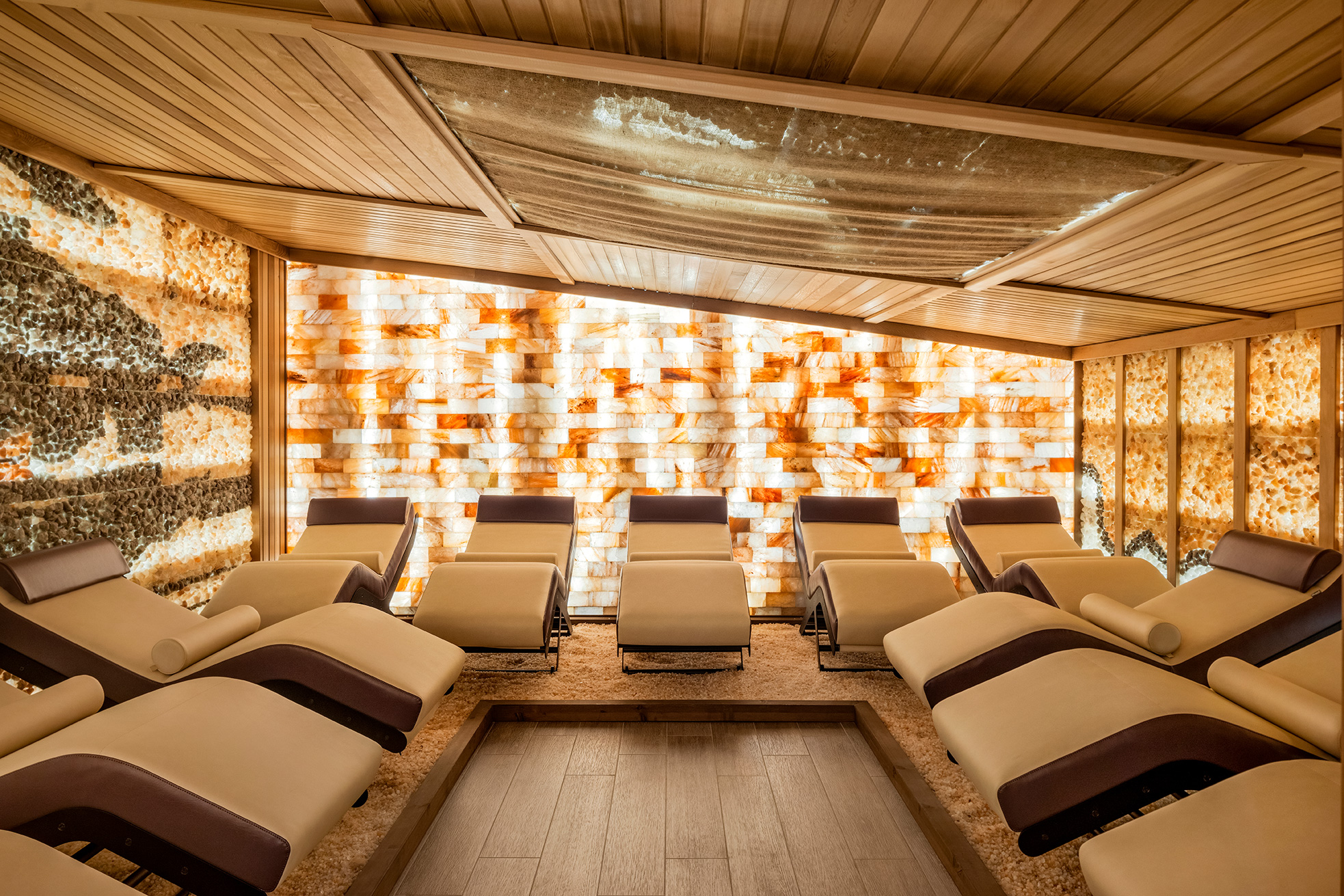 A Himalayan salt room with multiple reclining spa loungers arranged neatly. The room features warm lighting, walls adorned with illuminated salt crystals, and a ceiling with wooden beams, creating a serene and peaceful atmosphere.