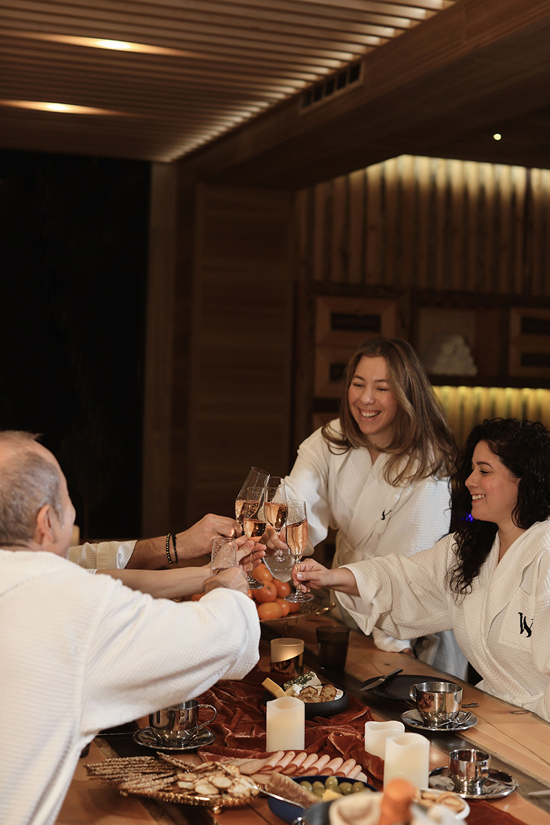 A group of people in white robes are cheerfully toasting with champagne glasses in a warm and cozy spa lounge. The wooden décor and soft lighting create an inviting and relaxed ambiance.