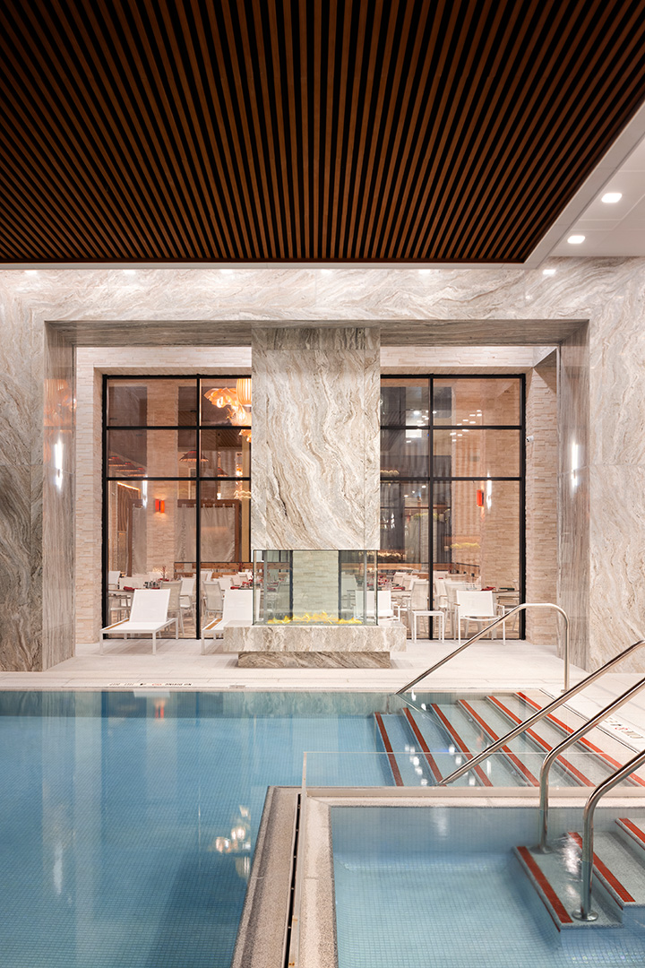 This is an indoor swimming pool area with a modern and luxurious design. The ceiling is adorned with parallel wooden slats over a reflective marble finish. The pool is bordered by elegant stone tiles, and there are steel railings leading into the water. In the background, through large glass windows, an intimate dining area with stylish furniture and warm lighting can be seen.