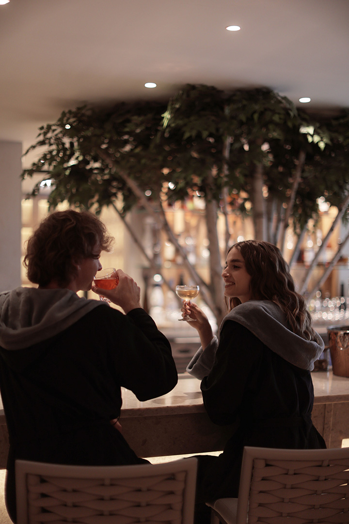 Two people in robes toasting with drinks in a cozy spa setting.