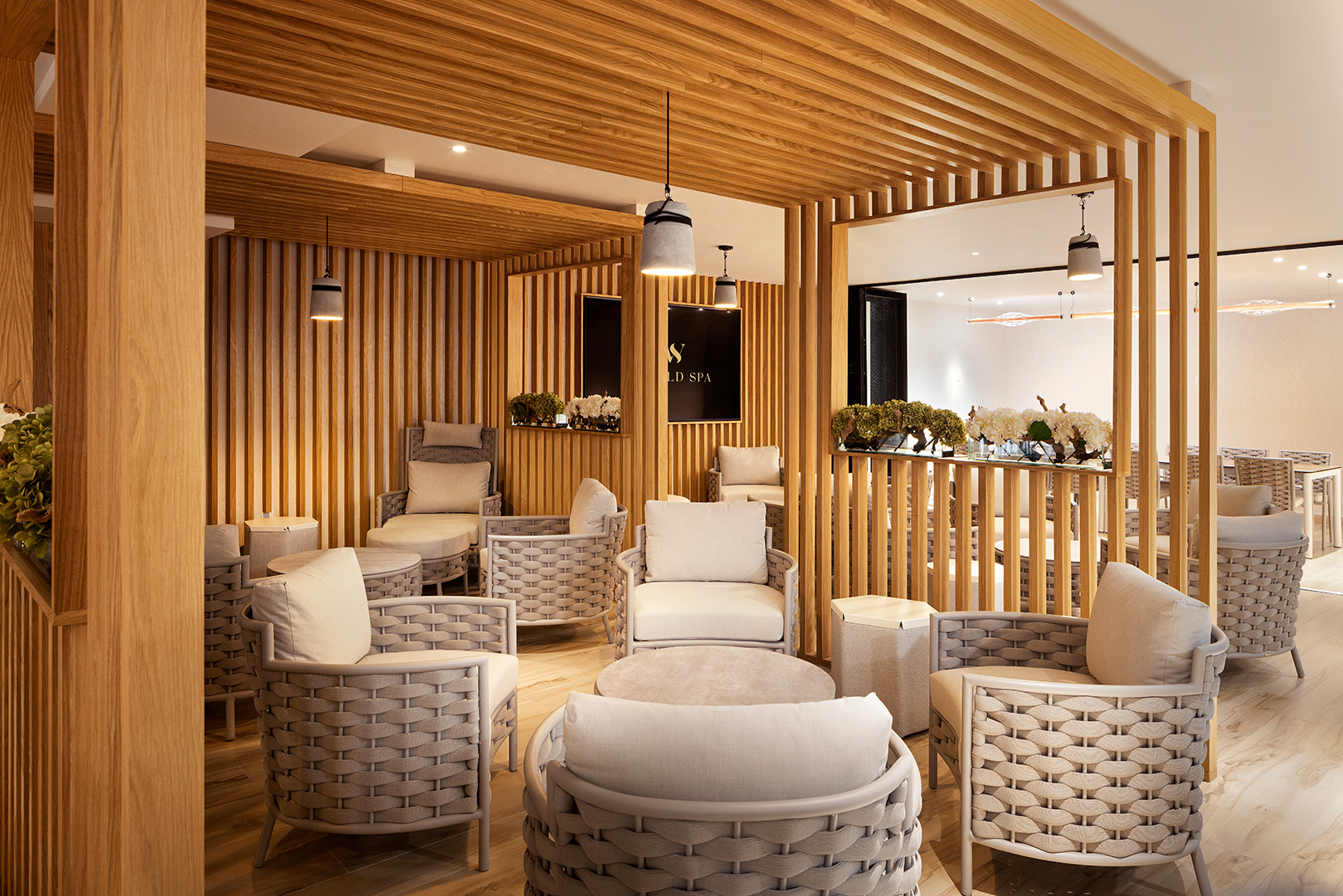 A luxurious lounge space boasting a contemporary design, with an array of woven chairs arranged for intimate conversations, complemented by soft pillows. The room features wooden slatted partitions, stylish pendant lights, and elegant floral decorations