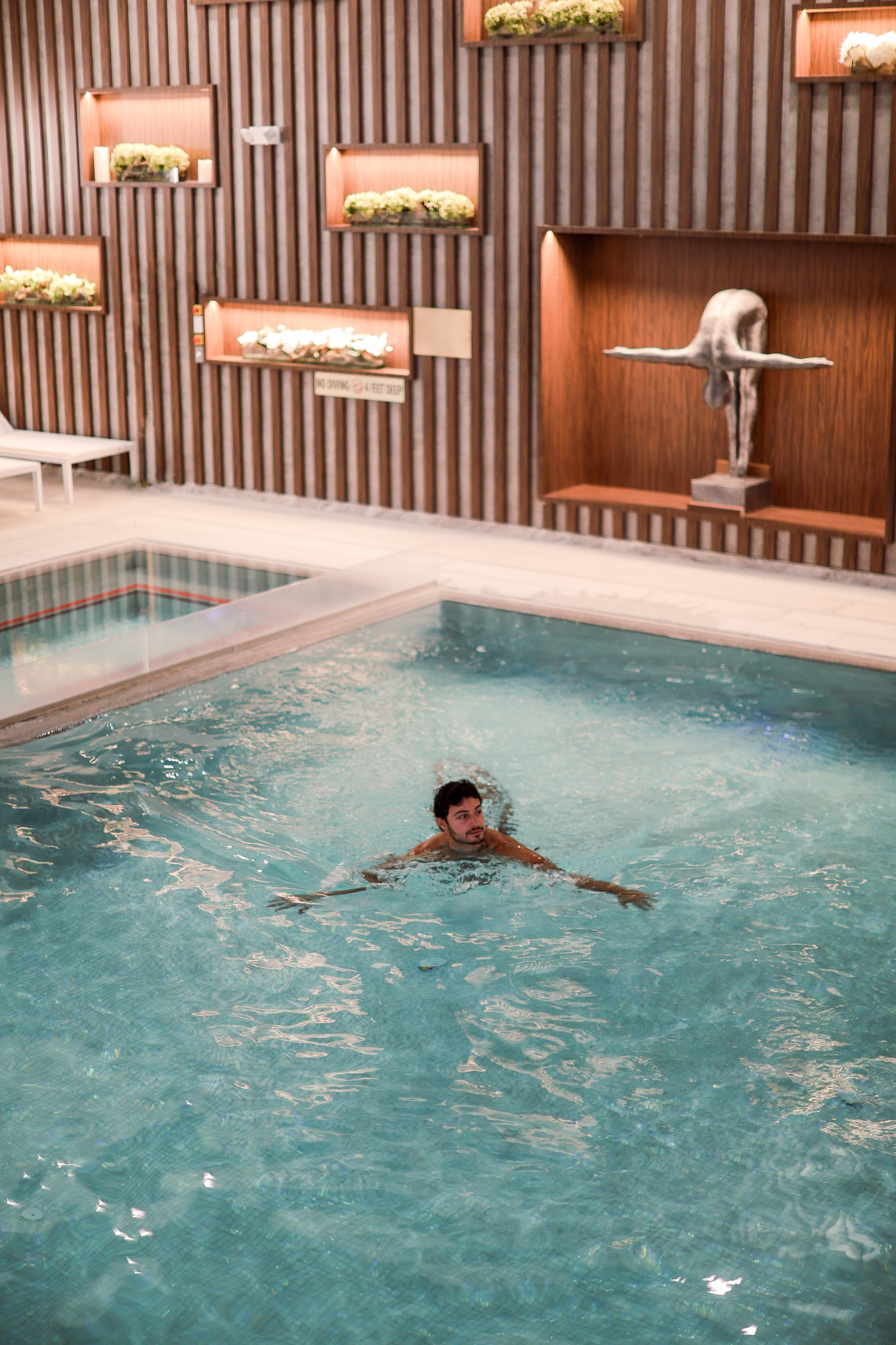 A person is floating in a clear, blue swimming pool. They have a relaxed posture with arms spread out. The pool is indoors, and the surrounding area features wooden walls with decorative plants and a sculpture. There is a sign on the wall indicating 'No Diving, Shallow Water