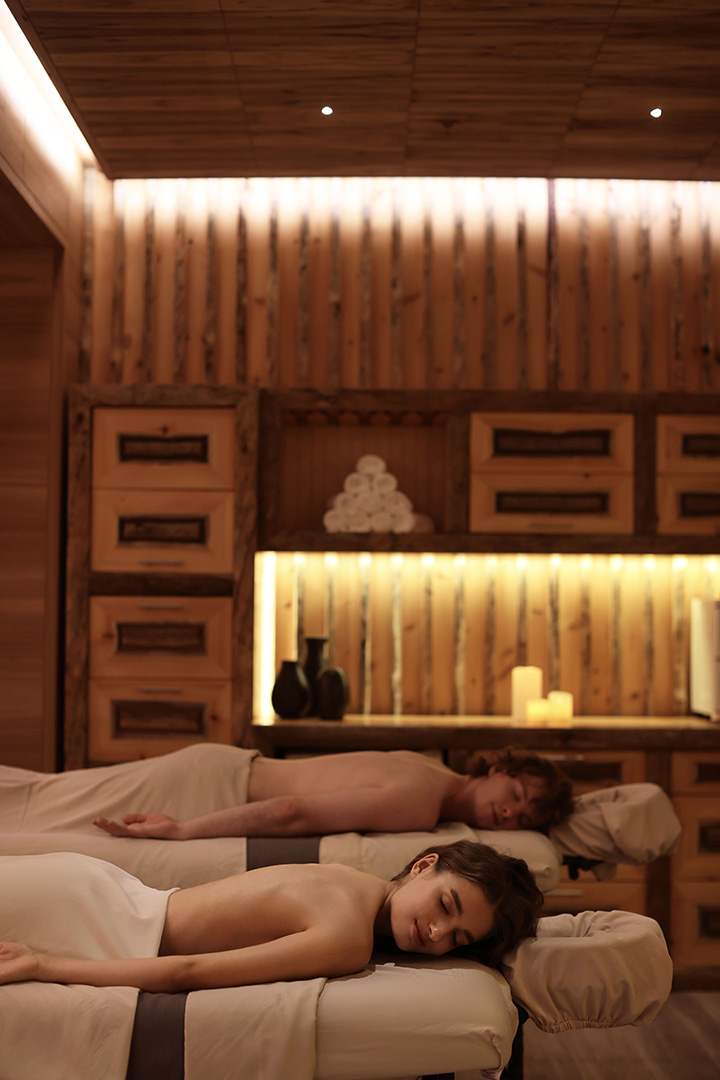 Two people are lying down on massage tables, covered by towels, in a tranquil and dimly lit spa room with wooden walls and soft ambient lighting, appearing very relaxed.